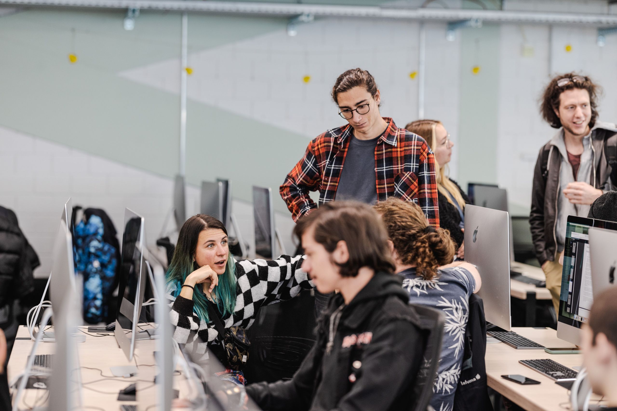 Eine Gruppe Menschen steht um einen Arbeitstisch in einem Büro mit vielen Laptops und diskutiert.