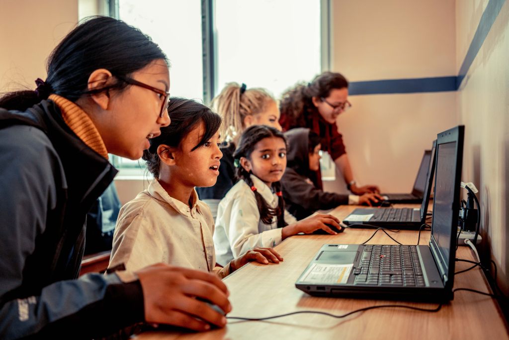 Indisch aussehende Mädchen sitzen in einem SChulzimmer an Tischen und bedienen Laptops.